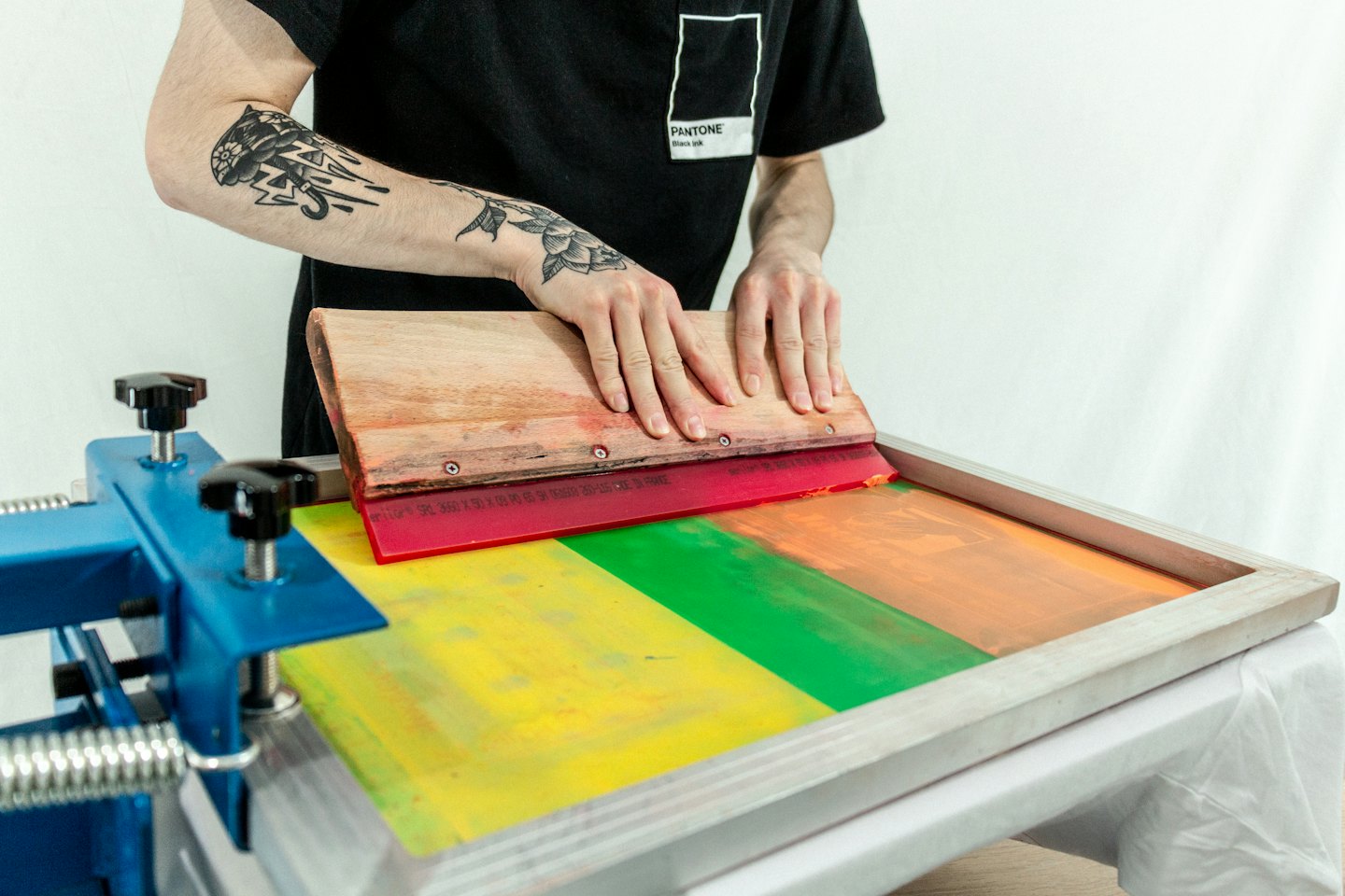 Manos con tatuajes pasan una rasqueta por una prensa de serigrafía.